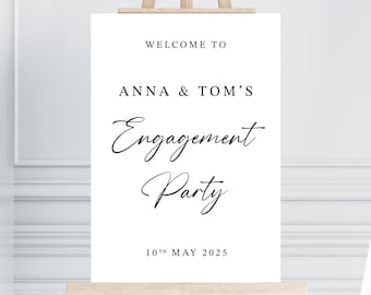 May include: A white sign on a wooden easel with the text "WELCOME TO ANNA & TOM'S ENGAGEMENT PARTY" in black script. The date "10TH MAY 2025" is printed at the bottom. The background is a light wall.