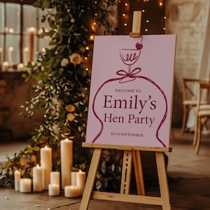 Könnte beinhalten: Ein hellrosa Schild auf einer Holzstaffelei, das die Gäste zu "Emily's Hen Party" mit einer Weinglas-Illustration willkommen heißt. Das Schild enthält die Daten "10-14 September". Der Hintergrund zeigt Kerzen, Grün und Lichterketten und schafft eine warme Atmosphäre.