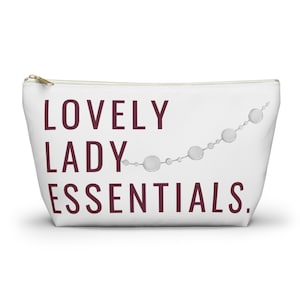 May include: White cosmetic bag with a gold zipper and the text "LOVELY LADY ESSENTIALS" in burgundy. A string of silver-colored circles decorates the upper right corner. This bag is designed to hold beauty products.