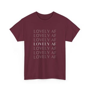 May include: A maroon t-shirt featuring the text "LOVELY AF" repeated vertically. The text is in white, in a simple font. The shirt has a crew neck and short sleeves, suitable for casual wear.