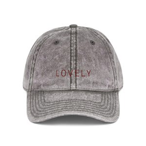 Peut inclure: Casquette de baseball grise avec le mot "LOVELY" brodé en rouge. La casquette a une visière incurvée et un bouton sur le dessus. Le tissu a un aspect délavé et vintage.