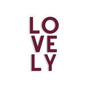 May include: A white sticker with the word "LOVELY" in a burgundy font. The letters are stacked vertically, with a white outline. The sticker is a simple, yet eye-catching design.