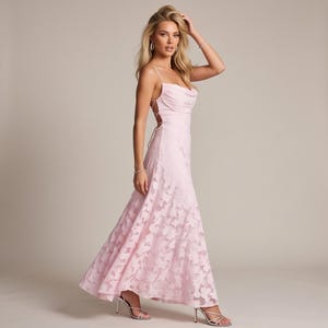 Könnte beinhalten: Ein bodenlanges, hellrosa Kleid mit Wasserfall-Ausschnitt, dünnen Spaghettiträgern und einem durchgehenden, spitzenartigen Blumenmuster. Das Kleid verfügt über eine seitliche Schnürung mit dünnen Bändern und einen fließenden Rock.