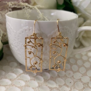 May include: A pair of gold-tone dangle earrings with a delicate, intricate design. The earrings feature a rectangular frame with a floral pattern.