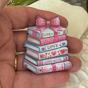 Pode incluir: Uma pilha de livros decorativos com a palavra "LOVE" nas lombadas, encimada por um laço rosa. Os livros são em tons de rosa e azul, decorados com corações e padrões florais. Pequeno objeto decorativo.