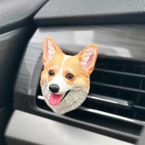 Peut inclure: Un clip de ventilation de voiture en forme de chien corgi. La tête du chien est orange, blanche et beige avec une langue rose. Le clip est fixé à une grille d'aération de voiture, avec un intérieur noir et argent.