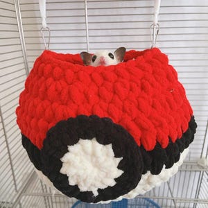 May include: A red, black, and white plush pet bed shaped like a ball, hanging inside a cage. The pet bed has a round shape with a black and white design on the bottom. A small, white animal with dark ears peeks out from the top opening.
