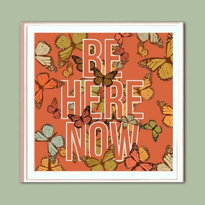 Op de afbeelding: Een perzikkleurige poster met een witte rand en een patroon van kleurrijke vlinders. De tekst "BE HERE NOW" is in wit op de poster gedrukt.
