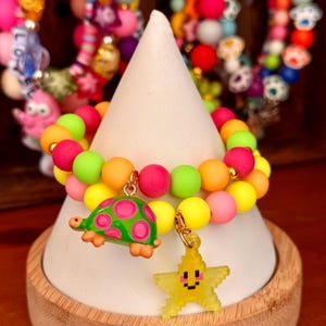 Puede incluir: Una colorida pulsera de cuentas con un dije de tortuga y estrella. La pulsera presenta cuentas vibrantes en tonos de rosa, amarillo, verde y naranja. El dije de tortuga es verde con lunares rosas, y el dije de estrella es amarillo.