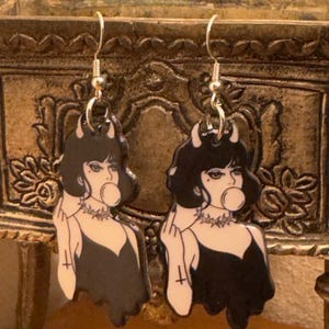 Puede incluir: Un par de pendientes colgantes plateados. Cada pendiente presenta una ilustración en blanco y negro de una mujer con cuernos, soplando una burbuja y vistiendo un vestido negro. Los pendientes tienen un gancho plateado.