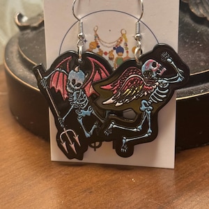 Puede incluir: Pendientes negros de Halloween con un esqueleto de diablo con un tridente y un esqueleto de ángel con alas. Los esqueletos tienen detalles en azul, rosa y amarillo. Los pendientes cuelgan de ganchos plateados.