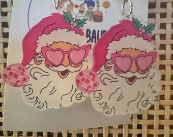 May include: Novelty earrings with Santa Claus faces wearing pink heart-shaped sunglasses and hats. The earrings have silver-coloured fish hook ear wires and disco ball-style pom-poms. The background includes the text "BAU".