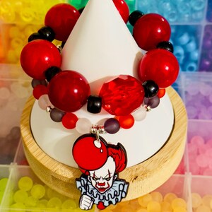 Puede incluir: Una pulsera de cuentas rojas y negras, con una gema roja facetada y un dije de payaso. El dije muestra un globo rojo y la cara del payaso. La pulsera se exhibe sobre un cono blanco.