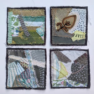 May include: Four square fabric coasters featuring layered textile scraps in shades of olive green, grey, and white. Each piece displays intricate hand-stitched embroidery, visible frayed edges, and a mix of floral and geometric patterns with white thread.