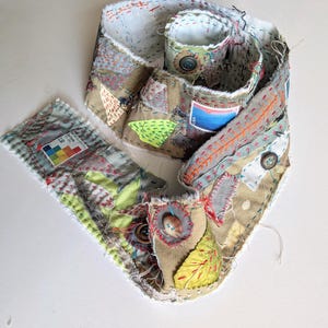 May include: A long, coiled fabric strip featuring patchwork construction with various textile scraps in shades of tan, green, and grey. It includes visible hand-stitched embroidery, several round buttons, and a blue postage stamp with the text USA.