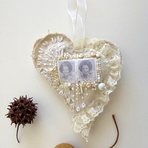May include: A heart-shaped fabric ornament with white lace trim, decorated with white beads, buttons, and two vintage postage stamps featuring black and white portraits. The ornament is hanging from a white ribbon.