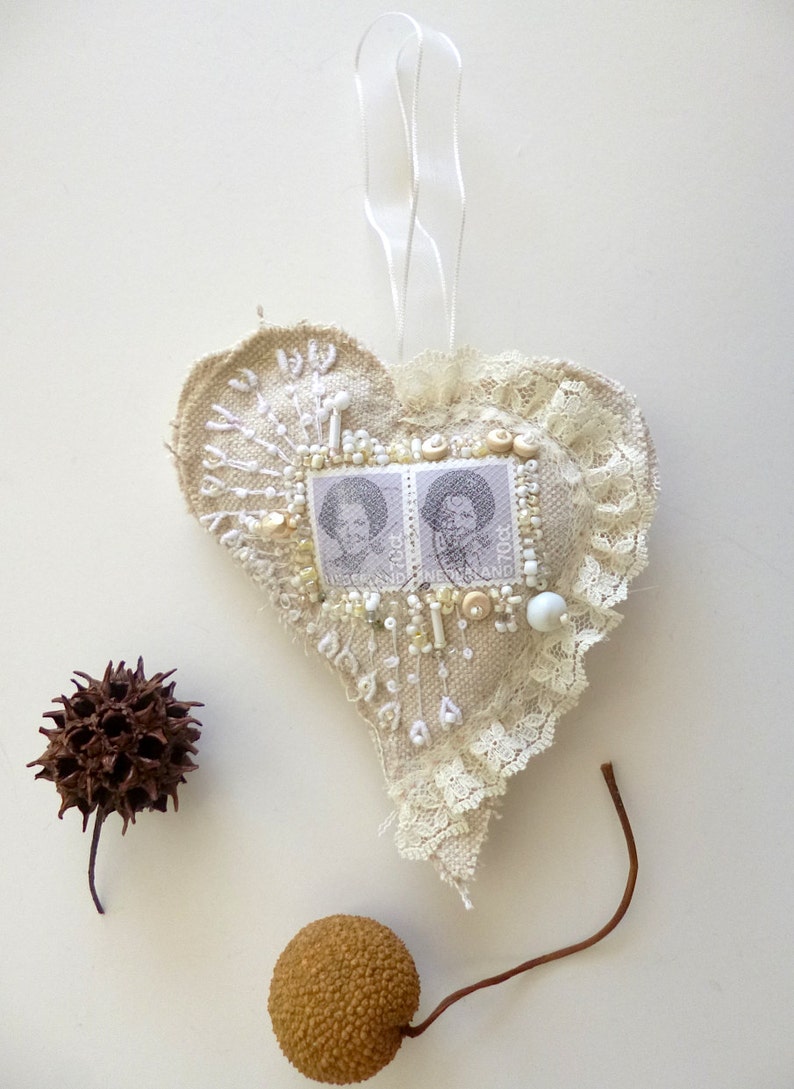 May include: A heart-shaped fabric ornament with white lace trim, decorated with white beads, buttons, and two vintage postage stamps featuring black and white portraits. The ornament is hanging from a white ribbon.