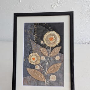 May include: A framed textile art piece featuring a dark gray denim background with layered fabric flowers in beige and cream, accented by bright orange centers. The design includes embroidered stems and leaves in light thread on a dark fabric base.