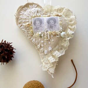 May include: A heart-shaped fabric ornament with white lace trim, decorated with white beads, gold sequins, and two vintage postage stamps featuring portraits of women. The ornament is hanging from a white ribbon.
