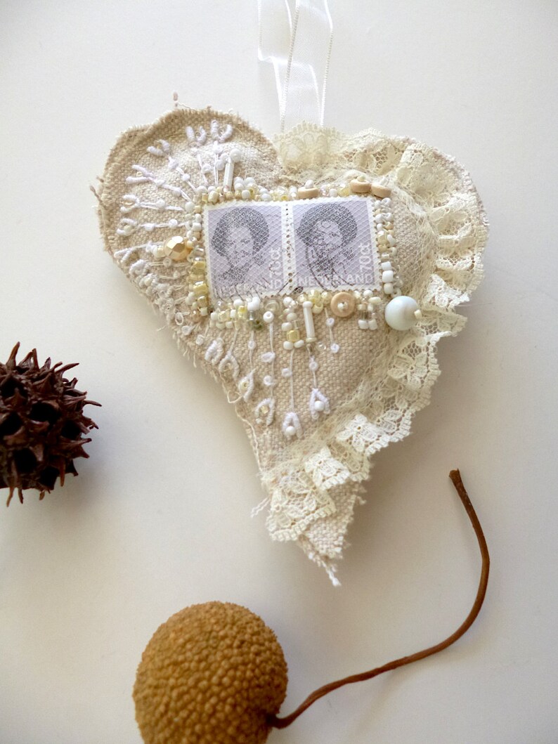 May include: A heart-shaped fabric ornament with white lace trim, decorated with white beads, gold sequins, and two vintage postage stamps featuring portraits of women. The ornament is hanging from a white ribbon.