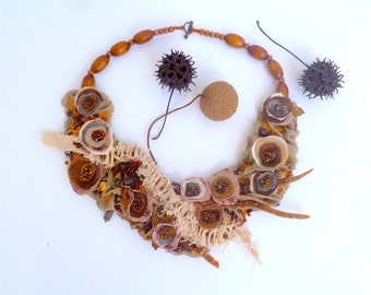 May include: A handcrafted necklace featuring a bib-style design with fabric flowers in shades of brown, beige, and orange. The necklace includes wooden beads and is accented with small, colorful beads. The design incorporates natural elements.