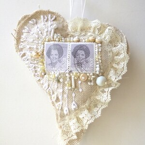 May include: A heart-shaped ornament made of burlap and lace, decorated with white beads, buttons, and two vintage postage stamps. The stamps feature a black and white image of a woman's face.