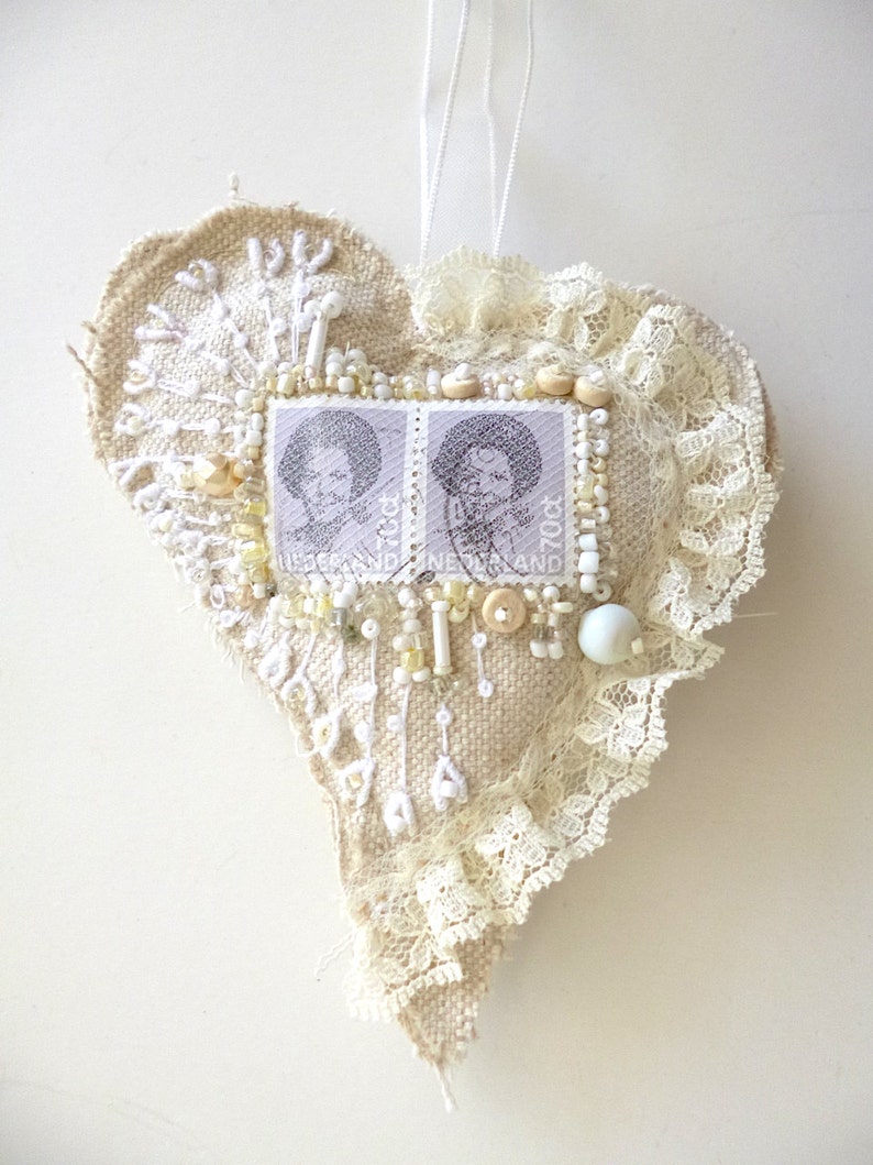 May include: A heart-shaped ornament made of burlap and lace, decorated with white beads, buttons, and two vintage postage stamps. The stamps feature a black and white image of a woman's face.