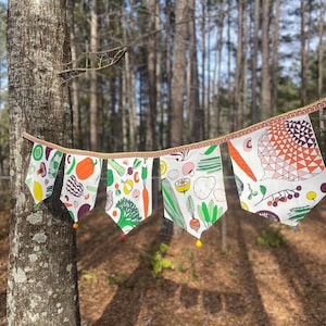 Könnte beinhalten: Ein farbenfrohes Banner mit Gemüseillustrationen, darunter Karotten, Brokkoli und Tomaten, hängt an einem Baum. Das Banner hat eine dekorative Bordüre und gelbe Pompons. Der Hintergrund ist ein Wald mit hohen Bäumen und Sonnenlicht.