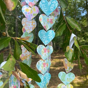 May include: A garland of paper hearts in various pastel colors, including blue, pink, and yellow, hangs from a tree branch. Each heart features a unique floral or patterned design. The background shows a blurred view of a green forest.