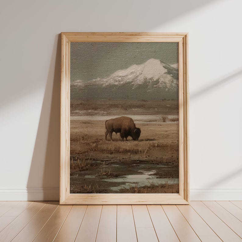 May include: A framed landscape print featuring a bison grazing in a field with a snow-capped mountain in the background. The artwork has a muted color palette of browns, greens, and grays, with a natural wood frame.