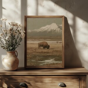 May include: A framed landscape print featuring a bison grazing in a field with a snow-capped mountain in the background. The artwork has a vintage aesthetic with muted earth tones. The frame is made of wood. A vase of flowers sits on a wooden surface.