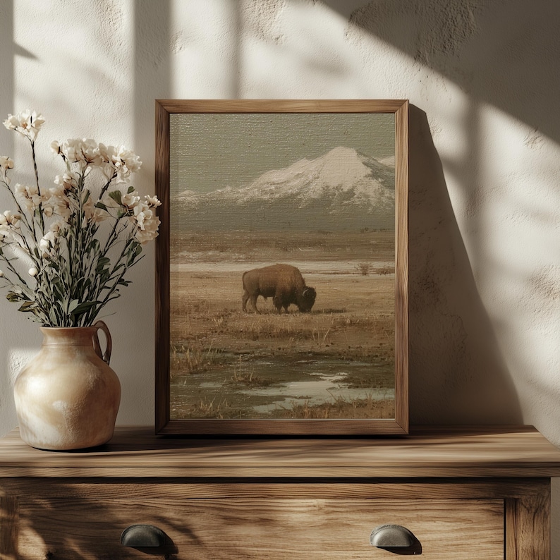 May include: A framed landscape print featuring a bison grazing in a field with a snow-capped mountain in the background. The artwork has a vintage aesthetic with muted earth tones. The frame is made of wood. A vase of flowers sits on a wooden surface.