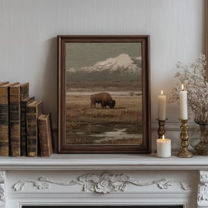 May include: A framed landscape painting depicts a bison grazing in a field with a snow-capped mountain in the background. The painting is in a dark wooden frame, displayed on a white mantelpiece with candles and antique books.