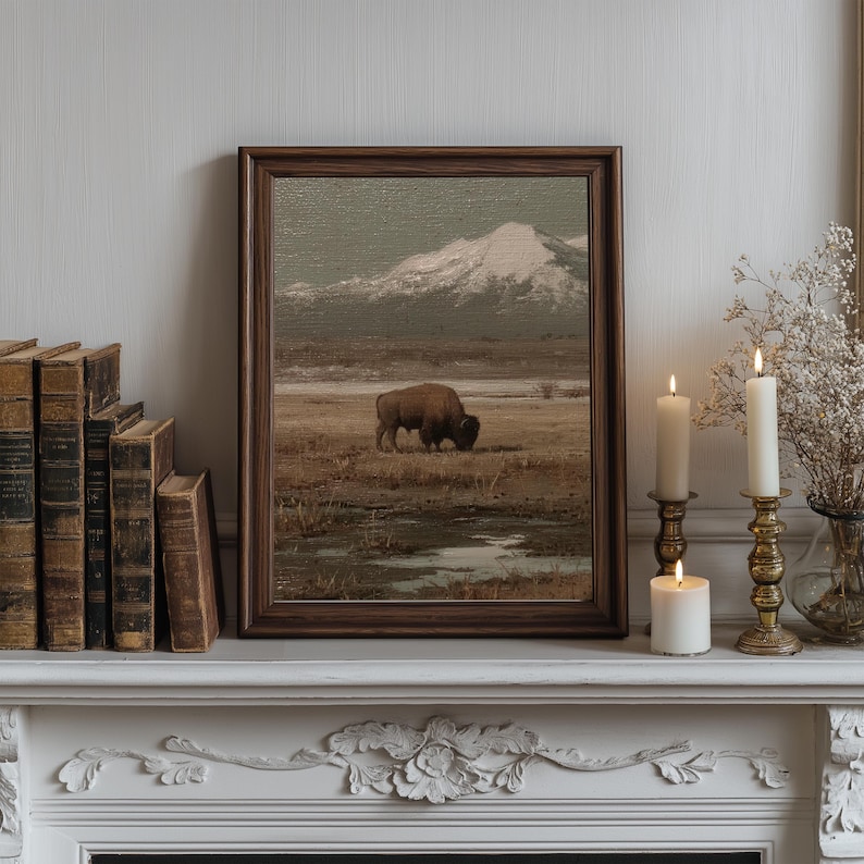 May include: A framed landscape painting depicts a bison grazing in a field with a snow-capped mountain in the background. The painting is in a dark wooden frame, displayed on a white mantelpiece with candles and antique books.