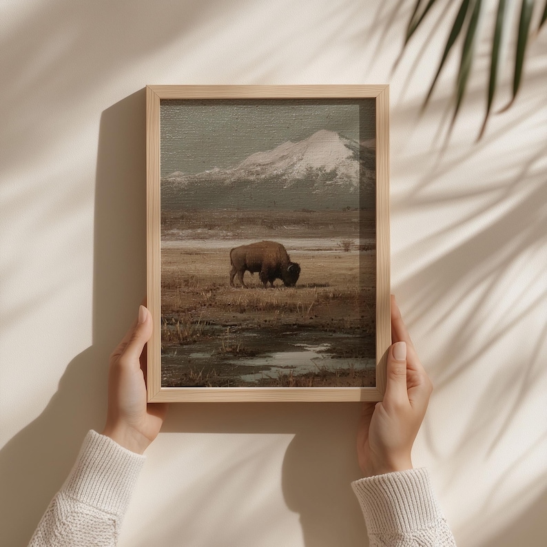May include: A framed landscape print featuring a bison grazing in a field with a snow-capped mountain in the background. The artwork has a muted color palette of browns, greens, and grays, creating a serene and naturalistic scene.
