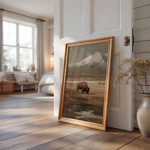 May include: A framed landscape painting featuring a bison grazing in a field with a snow-capped mountain in the background. The artwork has a gold-colored frame and is leaning against a white door in a room with hardwood floors.