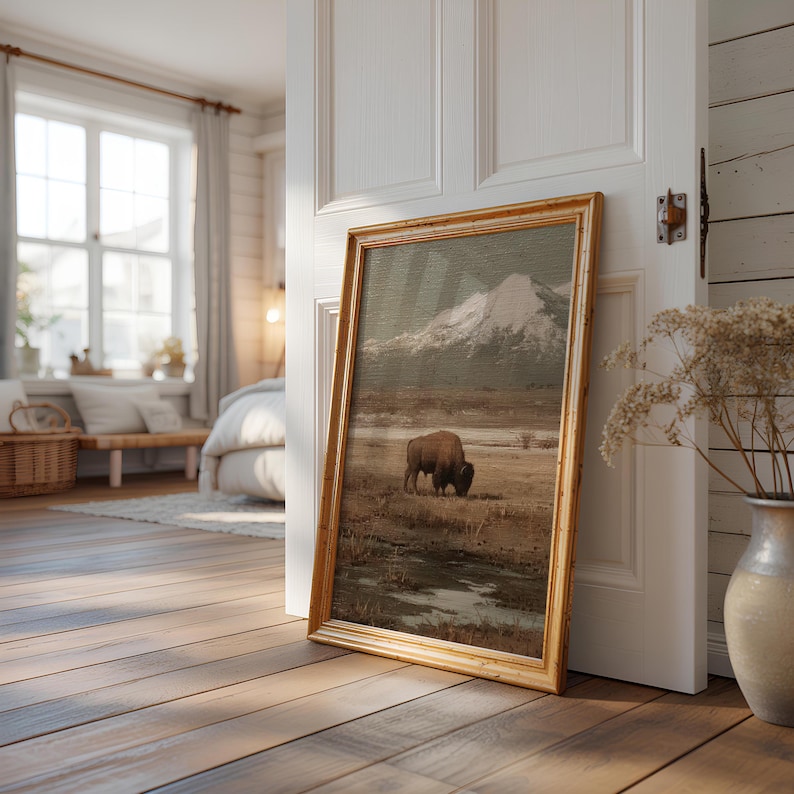 May include: A framed landscape painting featuring a bison grazing in a field with a snow-capped mountain in the background. The artwork has a gold-colored frame and is leaning against a white door in a room with hardwood floors.