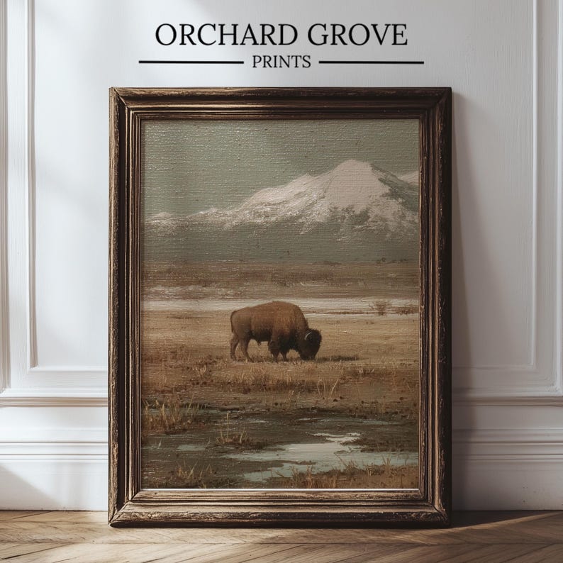 May include: Framed print featuring a vintage landscape painting. The artwork depicts a brown bison grazing in a field with a snow-capped mountain in the background. The print is in a dark brown wooden frame. The text "ORCHARD GROVE PRINTS" is at the top.