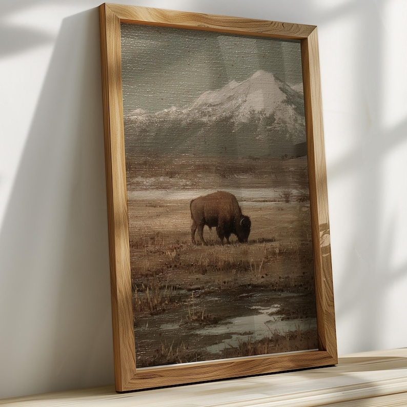 May include: A framed landscape print featuring a bison grazing in a field with a snow-capped mountain in the background. The artwork has a muted color palette of browns, greens, and grays, and is encased in a natural wood frame.