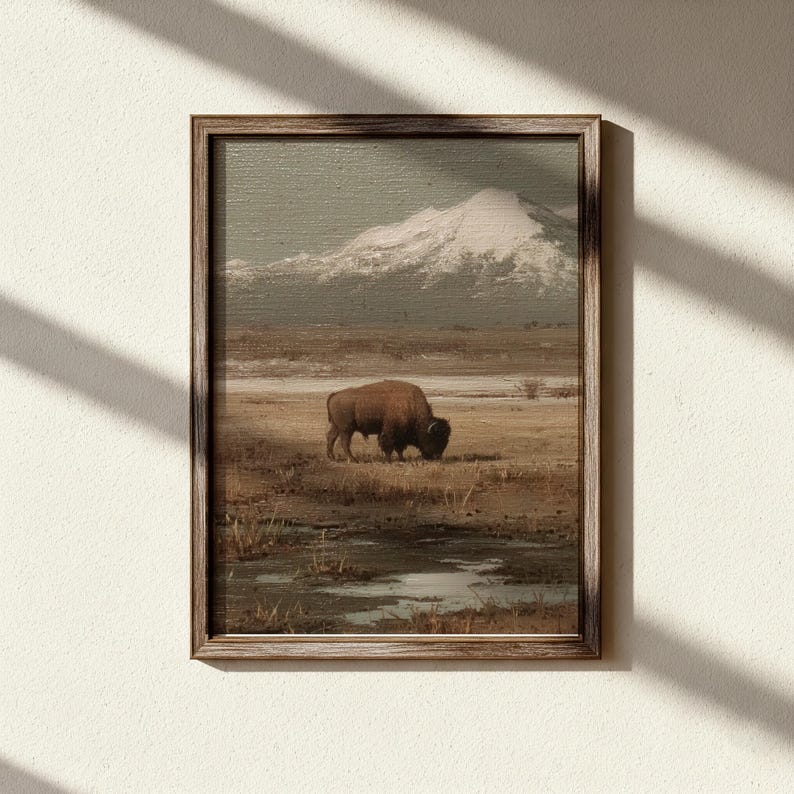 May include: Framed landscape art print featuring a bison grazing in a field with a snow-capped mountain in the background. The artwork has a vintage aesthetic with muted earth tones and a wooden frame.