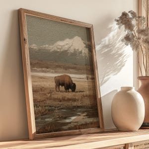 May include: A framed print depicts a bison grazing in a field with a mountain in the background. The artwork has a vintage aesthetic with muted tones of brown, beige, and gray. The wooden frame adds a rustic touch to the landscape scene.