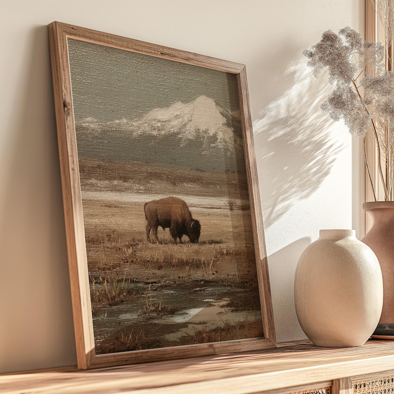 May include: A framed print depicts a bison grazing in a field with a mountain in the background. The artwork has a vintage aesthetic with muted tones of brown, beige, and gray. The wooden frame adds a rustic touch to the landscape scene.