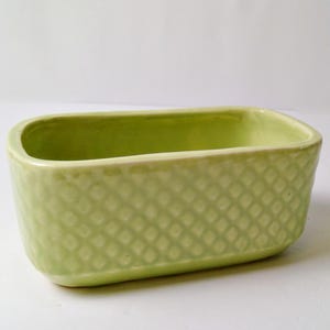May include: A light green rectangular ceramic container with a textured diamond pattern on the sides. The interior is a lighter shade of green. The container has rounded corners and a smooth rim.