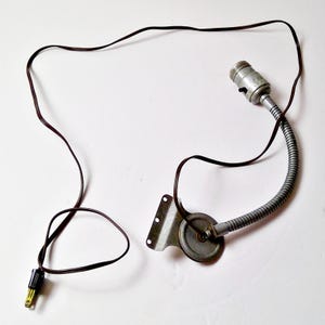May include: A vintage, flexible-neck lamp with a metal shade and a brown electrical cord. The lamp features a silver-colored, adjustable gooseneck and a standard light bulb socket. The cord has a two-prong plug.