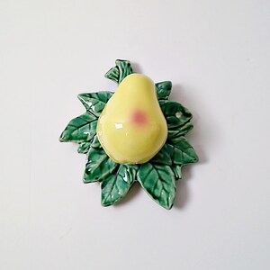 May include: A ceramic pear with a yellow glaze and a pink blush on the side, sitting on a bed of green leaves.