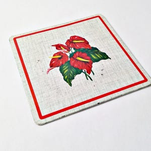 May include: Square metal trivet with a red border and a floral design. The trivet features a light gray background with a red and yellow anthurium flower design and green leaves.