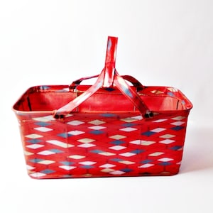 May include: A red metal picnic basket with a handle. The basket features a diamond pattern in white, blue, and yellow. The handle is also red with blue and white accents. The basket is rectangular and appears to be vintage.