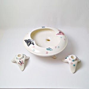 May include: A cream-colored ceramic ashtray and two matching candle holders. The ashtray is round with a central depression and three small feet. Each piece is decorated with painted butterflies in pink, blue, and black.