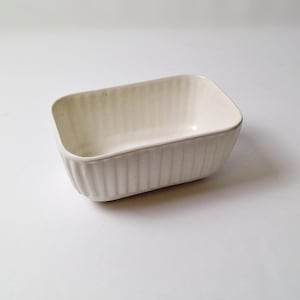 May include: A rectangular, off-white ceramic baking dish with vertical ribbed detailing on the sides. The dish has rounded corners and a smooth interior, suitable for baking and serving. The dish is empty and sits on a white surface.