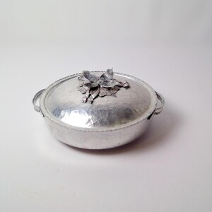 May include: A silver-colored, oval serving dish with a hammered texture and a decorative flower on the lid. The dish has two small handles and a beaded rim. A vintage piece for serving food.